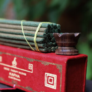 Zestaw kadzideł Kapha- grube kadzidła, podstawka i czerwone opakowanie kadzideł, leżą na stole na tle zieleni