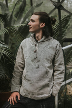 Mężczyzna z długimi włosami w kucyku stoi na tle zieleni w brązowej bluzie w jodełkę