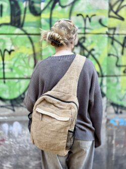 Kobieta stojąca tyłem ubrana w szary sweter na tle ściany z zielonym graffiti ma na sobie beżowy plecak wykoany z pokrzywy