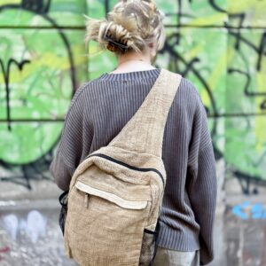 Kobieta stojąca tyłem ubrana w szary sweter na tle ściany z zielonym graffiti ma na sobie beżowy plecak wykoany z pokrzywy