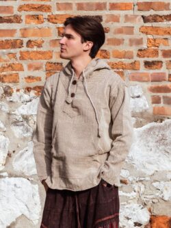 Mężczyzna ubrany w brązowe spodnie baggy i beżową bluzę w jodełkę, stoi na tle ceglanego muru