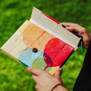 Osoba trzymająca kolorowy notes z papieru czerpanego z suszonymi liścmi i guziczkiem na okładce