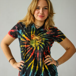 Osoba o blond włosach stoi na jednolitym tle, ubrana w kolorową koszulkę tie-dye z elfickim kapturem