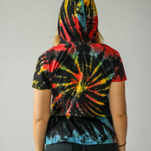 Osoba o blond włosach stoi na jednolitym tle, ubrana w kolorową koszulkę tie-dye