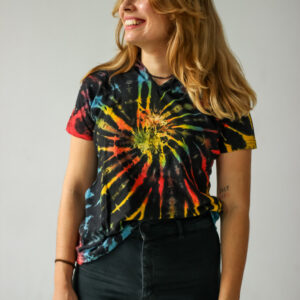 Osoba o blond włosach stoi na jednolitym tle, ubrana w kolorową koszulkę tie-dye z elfickim kapturem