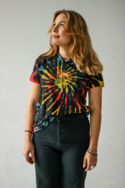 Osoba o blond włosach stoi na jednolitym tle, ubrana w kolorową koszulkę tie-dye