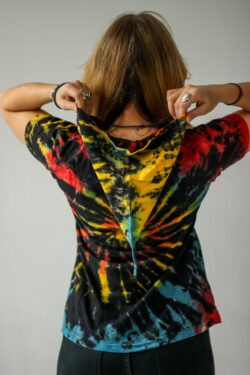 Osoba o blond włosach stoi na jednolitym tle, ubrana w kolorową koszulkę tie-dye z elfickim kapturem