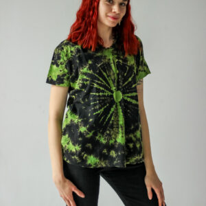 Osoba o rudych włosach stoi na jednolitym tle, ubrana w zieloną koszulkę tie-dye i czarne spodnie.