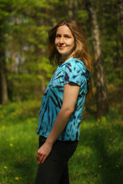 Osoba ubrana w niebieską bluzkę z kapturem z nadrukiem tie-dye stoi na zewnątrz, w tle widać drzewa.