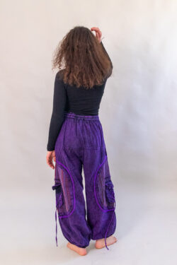 Kobieta w rozpuszczonych, kręconych włosach w czarnej bluzce i fioletowych baggy spodniach w vintage stylu z etnicznymi wzorami i kieszonkami po bokach.