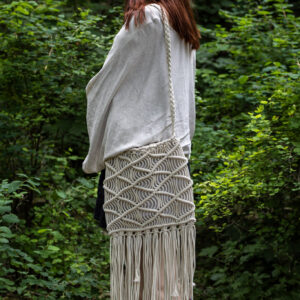 Kobieta w białej bluzce z beżową boho torebką z makramy z dużymi frędzlami stoi na tle zieleni