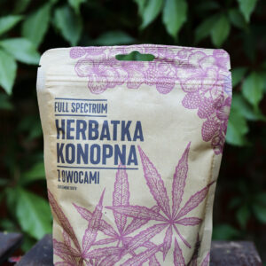 Opakowanie herbatki konopnej BIO z owocami stoi na drewnianym stoliku na tle zieleni.