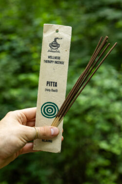 Osoba trzymająca opakowanie kadzideł Pitta oraz pałeczki kadzideł na tle zieleni.