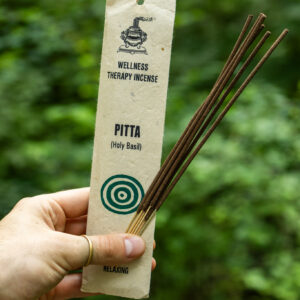 Osoba trzymająca opakowanie kadzideł Pitta oraz pałeczki kadzideł na tle zieleni.