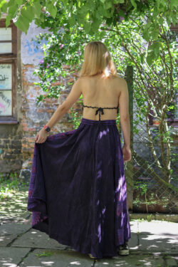 Kobieta w blond włosach i czarnym szydełkowym topie w fioletowej maxi spódnicy w mandale stoi tyłem na tle zieleni