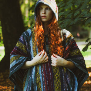 Kobieta w długich rudych, kręconych włosach ubrana w długie ponczo w etno wzory z kapturem, stoi na tle zieleni