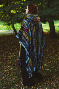 Kobieta w długich rudych, kręconych włosach ubrana w długie ponczo w etno wzory z kapturem, stoi na tle zieleni