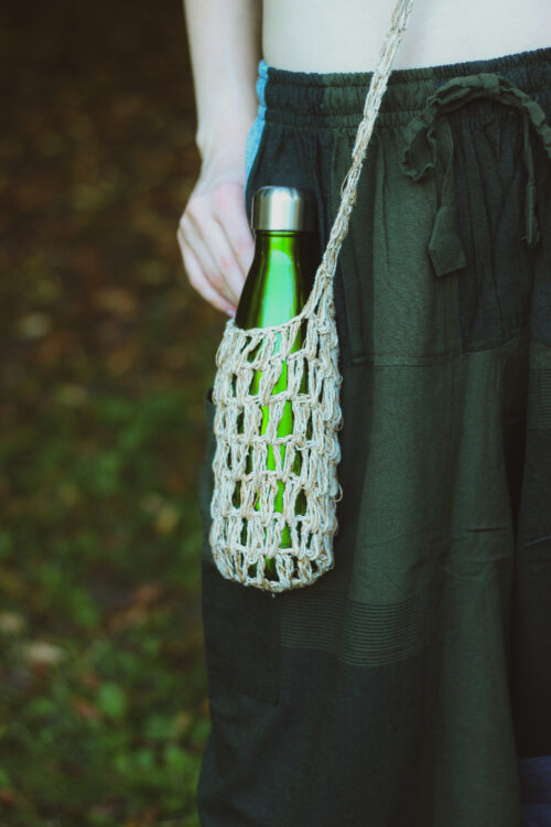 Kobieta w zielonych szarawarach ma przewieszone beżowe nosidełko na butelkę z konopi, a wnim zielony termos