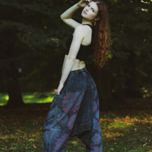 Kobieta w długich rudych włosach w czarnym topie i szarych szarawarach ze wzorem mandali stoi na tle zieleni