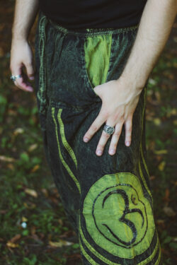 Mężczyzna ubrany w czarną bluzkę wkłada dłoń do kieszeni zielonych szarawarów z haftem z symbolem OM