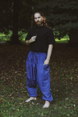 Mężczyzna ubrany w czarną koszulę z krótkim rękawem i niebieskie szarawary stoi boso na tle zieleni