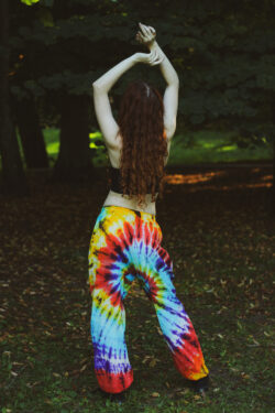 Kobieta w długich rudych włosach w czarnym topie i kolorowych spodniach z efektem tie-dye w hippie stylu stoi na tle zieleni