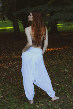 Kobieta z rudymi włosami ubrana w boho białe szarawary i beżowy top stoi boso na tle zieleni