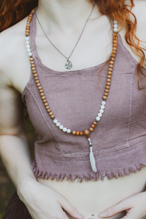 Mala z sandałowca Kobieta w długich, rudych kręconych włosach ubrana w beżowy boho top oraz drewniany naszyjnik - malę na tle zieleni