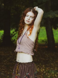 Kobieta w długich, rudych kręconych włosach ubrana w patchworkową spódnicę i beżowy boho top oraz fioletowy naszyjnik - malę stoi na tle zieleni