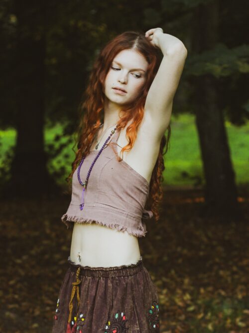 Boho top Kobieta w długich, rudych kręconych włosach ubrana w patchworkową spódnicę i beżowy boho top oraz fioletowy naszyjnik - malę stoi na tle zieleni