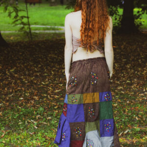 Kobieta w długich, rudych kręconych włosach ubrana w patchworkową spódnicę i beżowy boho top stoi na tle zieleni