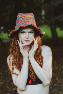 Kobieta z długimi rudymi włosami ubrana w czarny top i tęczowy kapelusz w skrzacim stylu