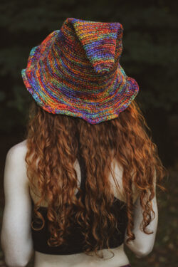 Kobieta z długimi rudymi wlosami stoi tyłem, ma na głowie tęczowy szpiczasty kapelusz w skrzacim stylu