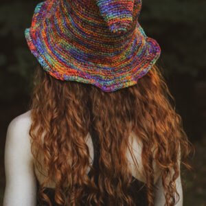 Kobieta z długimi rudymi wlosami stoi tyłem, ma na głowie tęczowy szpiczasty kapelusz w skrzacim stylu