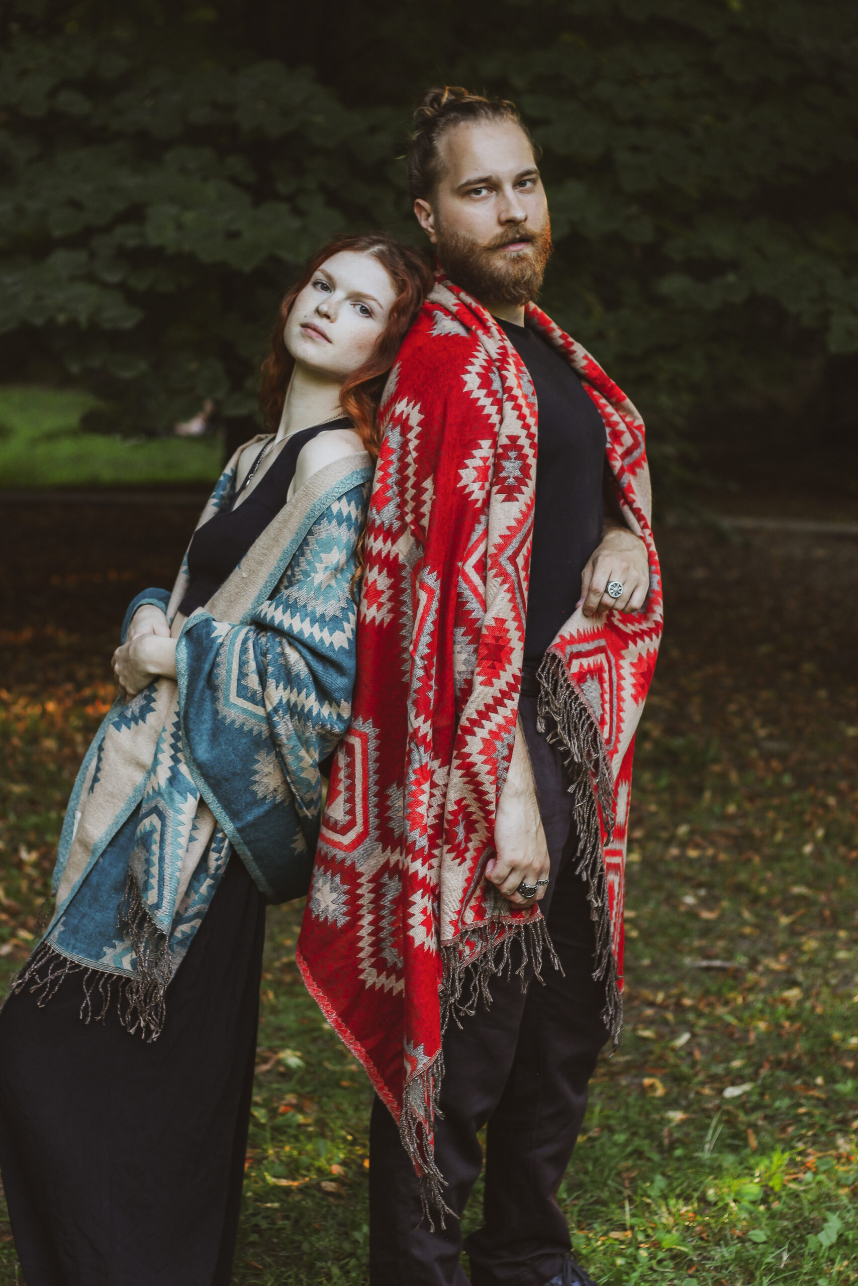 Kobieta i mężczyzna stoją oparci o siebie w parku ubrani w czarne ubrania i kolorowe szale w etno wzory