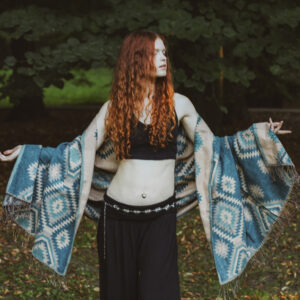Kobieta ubrana na czarno z długimi rudymi włosami stoi w parku owinięta w niebieski etno szal