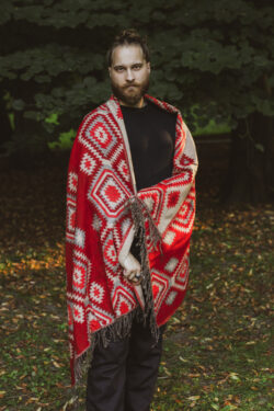 Meżczyzna ubrany na czarno stoi w parku owinięty w czerwony etno szal