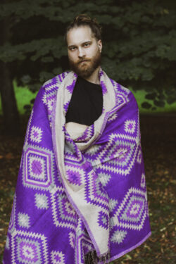 Meżczyzna ubrany na czarno stoi w parku owinięty w fioletowy etno szal
