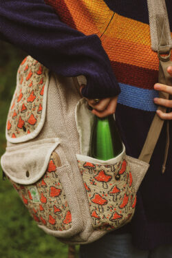 Kobieta w sweterku w paski ma stoi bokiem z dużym plecakiem w kolorze beżowym z nadrukami muchomorków i wyjmuje z plecaka butelkę