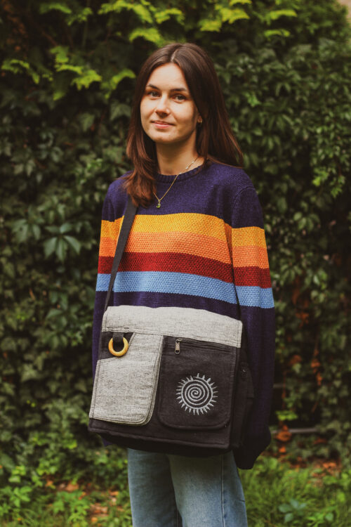 Kobieta w rozpuszczonych włosach i swetrze w paski z szaro-czarną torebką A4 ze spiralką stoi na tle zieleni