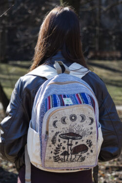 Osoba w czarnej skórzanej kurtce stoi tyłem i ma na plecach beżowo-niebiesko-czerwony plecak z ilustracją brązowych grzybów i roślin, wykonany z beżowej konopi i kolorowego materiału gheri, na tle parku