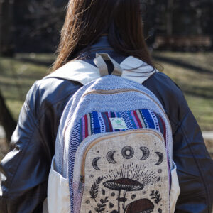 Osoba w czarnej skórzanej kurtce stoi tyłem i ma na plecach beżowo-niebiesko-czerwony plecak z ilustracją brązowych grzybów i roślin, wykonany z beżowej konopi i kolorowego materiału gheri, na tle parku