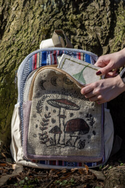 Osoba wkłada biały notes z zielonym liściem do przedniej kieszeni kolorowego plecaka z ilustracją brązowych grzybów, na tle drzewa