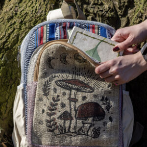 Osoba wkłada biały notes z zielonym liściem do przedniej kieszeni kolorowego plecaka z ilustracją brązowych grzybów, na tle drzewa