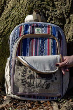 Osoba otwiera przednią kieszeń kolorowego plecaka z beżowymi kieszeniami po bokach i ilustracją brązowych grzybów, w środku znajduje się kolorowy materiał gheri, na tle drzewa