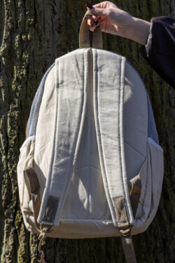 Osoba trzyma beżowy plecak z kieszeniami po bokach tyłem na tle drzewa