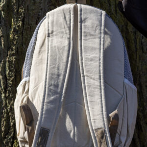 Osoba trzyma beżowy plecak z kieszeniami po bokach tyłem na tle drzewa