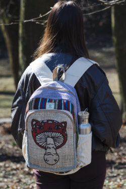 Osoba w czarnej skórzanej kurtce stoi tyłem w parku i ma na plecach beżowo fioletowo czerwony plecak z ilustracją muchomora z podobizną kota wykonany z beżowej konopi i kolorowego materiału gheri w bocznej kieszeni znajduje się srebrny bidon