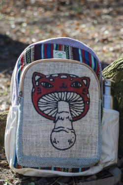 Kolorowy plecak leży na ziemi przy drzewie, ma beżowe kieszenie po bokach, jest wielkości A4 i wykonany z bawełny gheri oraz konopi, na przedniej kieszeni znajduje się ilustracja muchomora o podobiźnie kota
