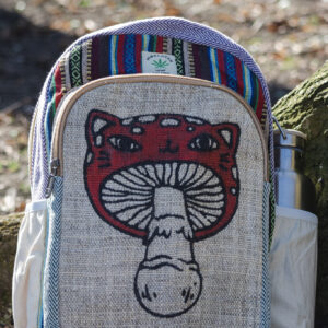 Kolorowy plecak leży na ziemi przy drzewie, ma beżowe kieszenie po bokach, jest wielkości A4 i wykonany z bawełny gheri oraz konopi, na przedniej kieszeni znajduje się ilustracja muchomora o podobiźnie kota